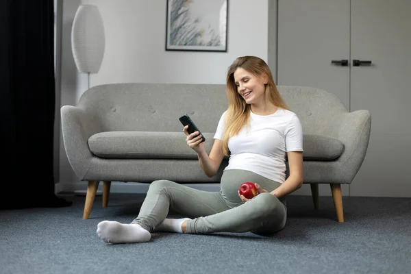 スマートフォンやリンゴを持っている妊婦を笑顔 自宅に座ってモバイルアプリのショッピングを使用して 妊娠概念 — ストック写真