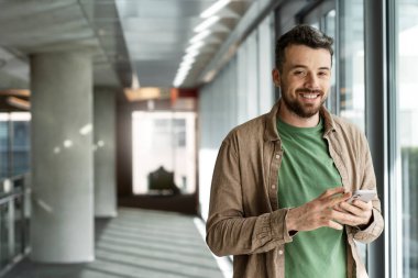 Genç Latin bir adam cep telefonu tutuyor, ofiste duruyor, gülümsüyor. İçerde akıllı telefon kullanarak günlük kıyafetler giyen şık bir adamın portresi.