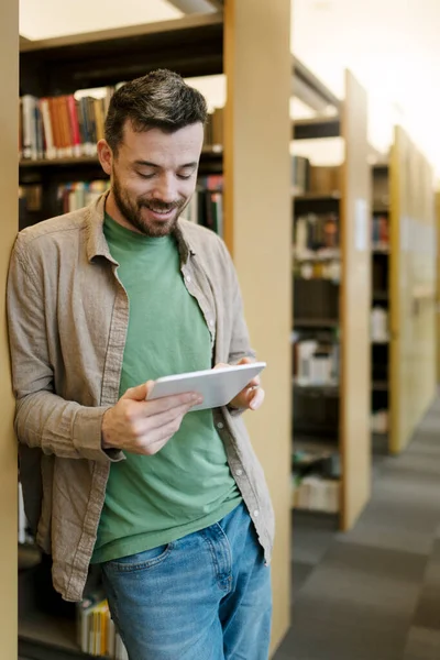 Attractive, bearded man is standing near book stand, using tablet, serching book. Middle aged hispanic student studying online in library, online education concept