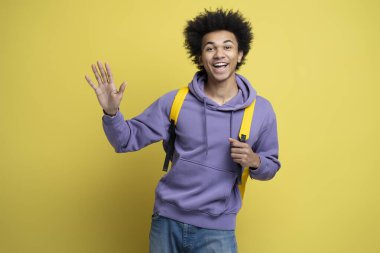 Smiling friendly African American man wearing stylish hoodie, waving hand isolated on yellow background. Portrait of happy successful university student looking at camera. Education concept 