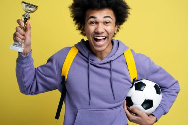 Young overjoyed African American man football player holding ball and trophy cup, celebration success isolated on yellow background. Sport victory, winning competition concept 