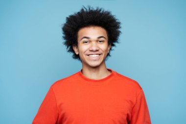 Mavi arka planda izole edilmiş şık günlük kıyafetler giyen kıvırcık saçlı, mutlu, Afro-Amerikan bir adam. Neşeli başarılı öğrenci kameraya bakıyor. Eğitim kavramı 