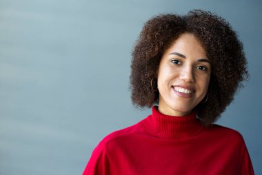 Kendine güvenen çok ırklı bir kadının yakın plan portresi, şık afro saç stili, kırmızı bir kazak giyiyor, kameraya kibarca gülümsüyor, gri bir duvar arkasında duruyor. - İnsanlar. Duygular