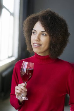 Güzel Afro-Amerikan genç kadın, gündelik giysiler içinde, şarap kadehi tutuyor lezzetli kırmızı şarap tadıyor, kendinden emin bir şekilde yan tarafa bakıyor. İçki endüstrisi. İçecek. Sommelier.