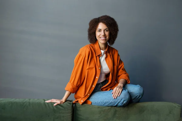 一个身穿橙色衬衫和蓝色牛仔裤 坐在客厅沙发上 面带微笑地看着相机的年轻而快乐的非洲裔美国女人自信的画像 人与生活方式概念 — 图库照片