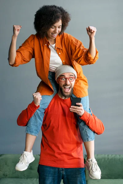 Overjoyed Caucasian young man in trendy glasses, carrying his girlfriend on his shoulders, holding smartphone, rejoicing and clenching fists from excitement while winning lottery, receiving great news