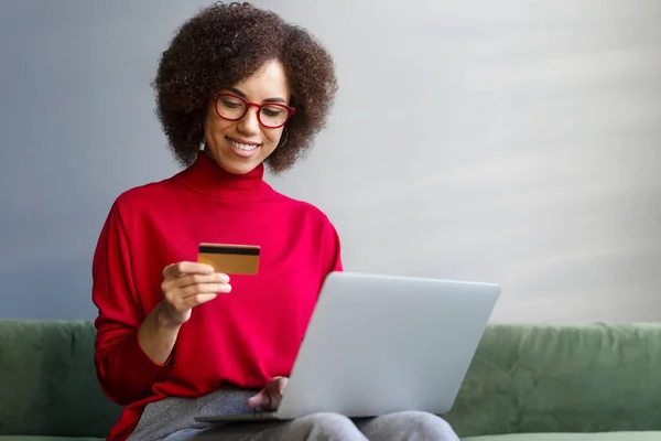 stock image Beautiful African American woman in casual clothes, sitting on sofa, holding credit card, using laptop shopping online, ordering food, booking, making cashless payment, transactions via mobile banking