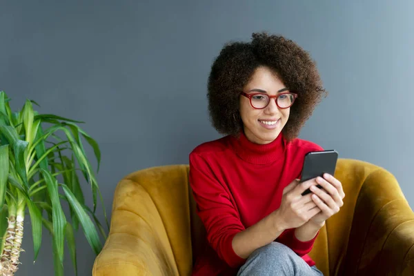 Smiling African American woman holding smartphone watching videos sitting on armchair at home. Happy modern female using mobile app shopping online, ordering food, reading text message, check email