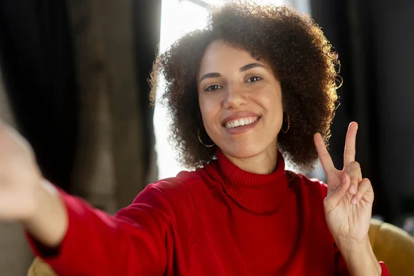 Smiling African American woman holding mobile phone , taking selfie, communication online, showing victory sign, having video call sitting at home. Happy influencer recording video using smartphone