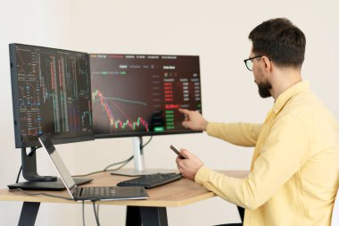 Ciddi bir tüccar cep telefonu tutuyor, dizüstü bilgisayar kullanıyor, bilgisayar monitörüne bakıyor kripto grafiği ile çevrimiçi çalışıyor, modern ofiste oturuyor, seçici odaklanıyor. Başarılı iş, çevrimiçi ticaret kavramı