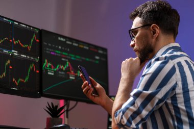 Cep telefonu tutan ciddi yakışıklı tüccarın gece portresi modern ofiste kripto grafiğiyle çalışan dizüstü bilgisayarını kullanıyor. Başarılı iş, çevrimiçi ticaret kavramı