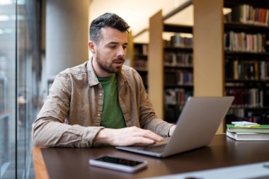 Modern kütüphanede oturan ciddi melez ırk öğrenimi, öğrenme, sınav hazırlığı, online eğitim kavramı. Yakışıklı orta yaşlı Latin metin yazarı. İş yerinde serbest çalışma projesi.