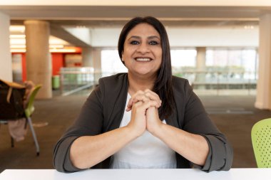 Olgun, kendine güvenen Asyalı iş kadını kameraya bakıp gülümsüyor, modern ofis içinde masa başında oturuyor, üretken ve başarılı bir iş gününden memnun görünüyor.