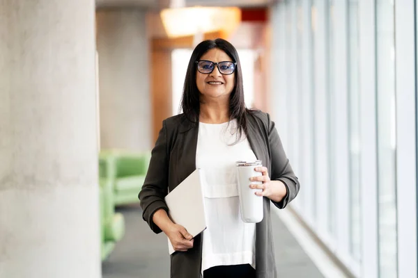 Confident mature Indian woman wearing eyeglasses holding digital tablet, bottle of water looking at camera in modern office. Portrait of successful smiling asian manager