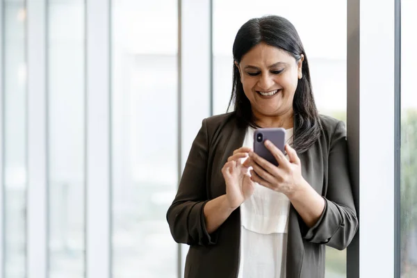 Happy smiling mature Asian woman holding mobile phone and checking social media content, scrolling news feed, standing bt windows in corporate office interior. Businesswoman using smartphone indoors