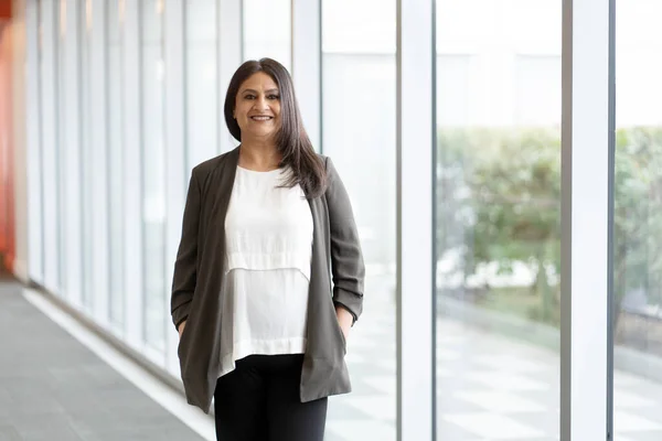 Confident mature Indian woman wearing stylish casual clothes, standing by large windows in the corporate office interior, smiling cutely looking at camera. Successful Asian people and business concept