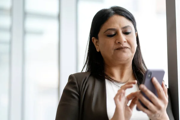 Confident mature Indian woman in formal suit, using mobile phone in corporate office, working online, browsing websites, text messaging, booking, ordering, planning business meeting with colleagues