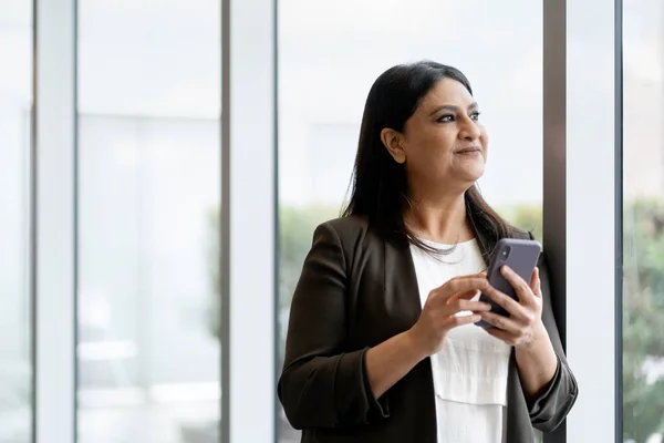 Close-up portrait of a charming mature Asian woman, wearing stylish casual clothes, standing by large windows in the corporate office interior with mobile phone in her hands People. Business concept