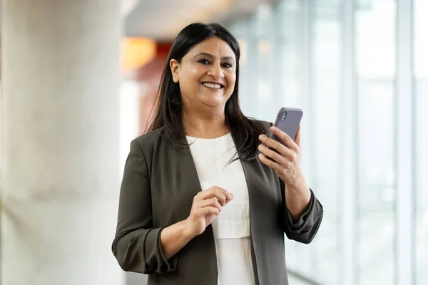 Smiling confident mature Asian woman in formal suit, holding mobile phone, looking at camera while standing in modern corporate office interior. People. Finance. Success. Business. Technology concept
