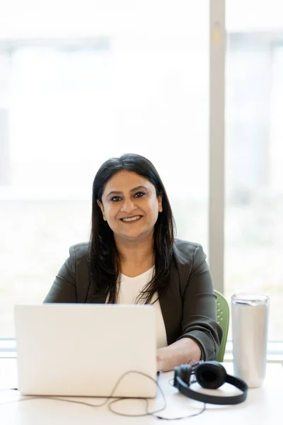 Confident portrait of a mature Indian woman in formal suit, sitting at desk with laptop, smiling a cheerful toothy smile and confidently looking at camera. Businesswoman at workplace in modern office