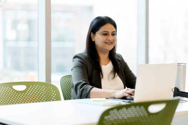 Confident positive Indian mature woman, a sales or executive manager using laptop, planning project, typing text on keyboard, browsing the internet, sitting at desk in modern corporate office