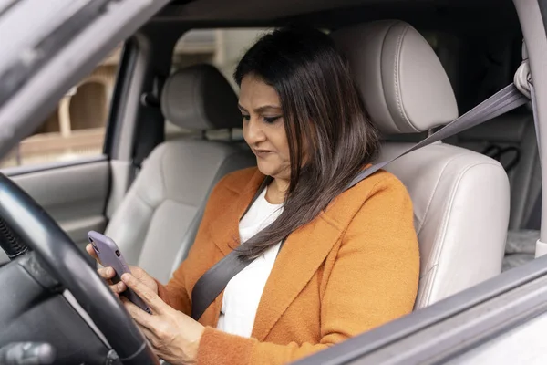 Confident multitasking mature female driver, businesswoman using smartphone, setting up navigator on mobile app, planning online meeting, sitting in driver\'s seat in car. People. Online communication