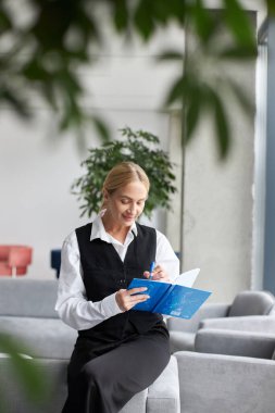 Çekici gülümseyen orta yaşlı iş kadını elinde defter tutuyor, planlama projesi yapıyor, modern ofiste çalışıyor. İş yerinde oturan başarılı yazarın portresi