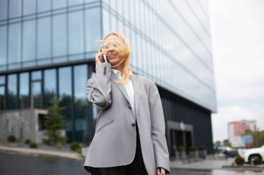 Cep telefonuyla konuşan bir kadın sokakta yürüyor, yüzünde kıllar olan güçlü bir rüzgar var. 