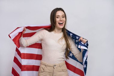 Güzel, vatansever, hayat dolu, genç bir kadın uzanmış ellerinde Amerikan bayrağıyla beyaz arka planda tek başına kameraya bakıp gülümsüyor.