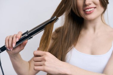 İzole beyaz arka planda saçları düzleştiren gülümseyen bir kadının elindeki modern elektrikli saç düzleştiriciye odaklan. Profesyonel saç şekillendirme aletleri. Kuaför. Berber. Saç bakımı