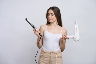 Kafkasyalı, kafası karışmış, kafası karışmış, saç düzleştirici ve kurutma makinesi tutan, beyaz arka planda izole edilmiş şık saç stili için hangi saç stili aletini kullanacağını seçen güzel bir kadın. Reklam alanı
