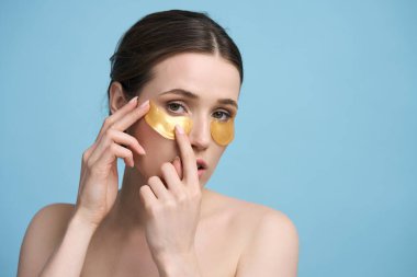 Sağlıklı, parlak ciltli güzel bir kadın, kameraya bakarken siyah halkaları çıkarmak için altın hidrojel bantlar takıyor ve gözlerinin altındaki şişkinliği yatıştırıyor, izole edilmiş mavi arka plan.