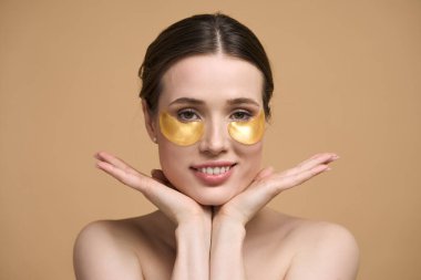 Yakından beyaz güzel bir kadının portresi altın hidrojeli anti pufiness bantları gözünün altında, ellerini yüzünün altında tutuyor, kameraya gülümsüyor, izole edilmiş bej arka plan