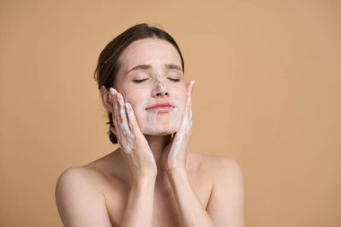 Çekici, beyaz bir kadın yüzüne sabun köpüğü sürüyor, masaj yapıyor, gözleri kapalı ve çıplak omuzlarıyla poz veriyor, bej arka planda izole edilmiş.