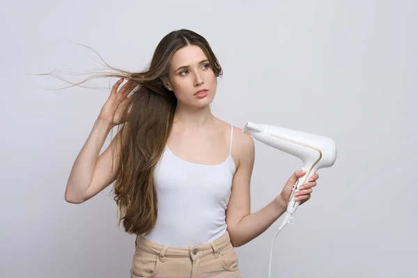 Beautiful Caucasian young woman with long silky glowing silky straight hair, dreamily looking aside, using hair dryer isolated white background. Hair care concept. People. Lifestyle. Morning routine