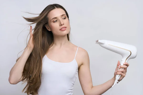 Isolated portrait on white background of Caucasian confident young pretty woman, wearing white top, holding stylish modern electric hair dryer and blowing warm air on her beautiful silky long wet hair