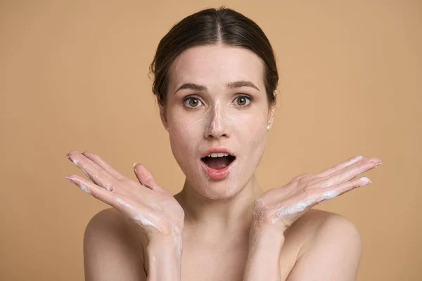 Amazed young woman with makeup remover cleansing foam on her face, looking at camera, posing with open mouth from surprise and bare shoulders, isolated on beige backdrop. Skin care and hygiene concept