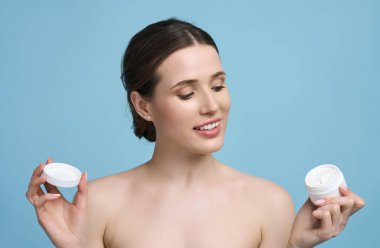 Gülümseyen beyaz kadın kozmetik maket kavanozu, yüz kremi kalıbı kavanozu tutuyor. Sağlıklı, parlak ciltli ve doğal makyajlı güzel bir kadın, izole edilmiş mavi arka planda çıplak omuzlar.