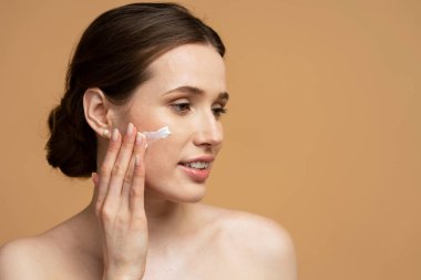 Yüzüne krem süren güzel, gülümseyen genç bir kadının yakın plan portresi. Bej arka planda izole edilmiş, kopyalama alanı. Cilt bakımı, doğal güzellik konsepti