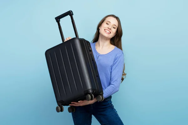 stock image Young smiling woman, traveler holding suitcase ready for vacation, isolated on blue background. Travel concept