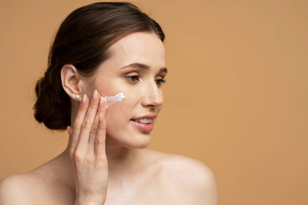 Closeup portrait of beautiful smiling young woman applying cream on face looking away isolated on beige background, copy space. Skin care, natural beauty concept