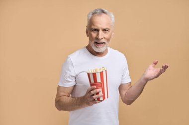 Beyaz tişörtlü neşeli bir son sınıf öğrencisi elinde patlamış mısırla tek kullanımlık bir kağıt kutu tutuyor, film, video, televizyon izlerken el kol hareketi yapıyor, bej arka planda izole edilmiş. Sinema Eğlence Hobisi