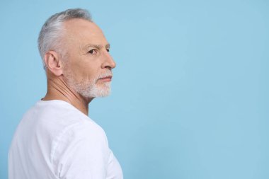 Beyaz saçlı, sakallı, yakışıklı, emekli, beyaz günlük tişörtlü, düşünceli, mavi arka planda soyutlanmış bir adamın yan portresi. Uzayı kopyala. İnsanlar