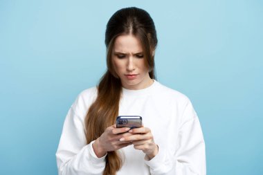 Beyaz tişört giyen, elinde akıllı telefon tutan, arkadaşlarıyla sohbet eden, emirler veren, konsantre bir yüz ifadesi olan ciddi düşünceli bir kadın. Mavi arka planda kapalı stüdyo çekimi 