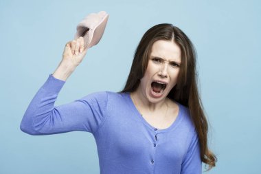 Ayakkabı tutan ve çığlık atan sıradan giysili esmer genç kadın portresi. İnsanlar duygularını ifade eder. Mavi arka planda kapalı stüdyo çekimi 