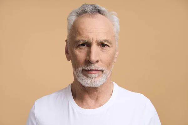 一个英俊的白种人 六十岁的白发苍苍的大胡子老人 身穿白色T恤 自信地看着相机 与米色背景隔离 人的情感生活方式 — 图库照片