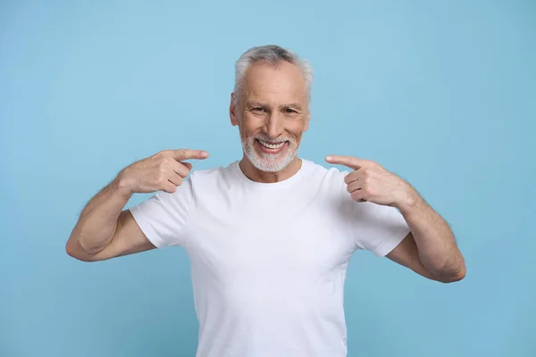 白种人白发苍苍的大胡子老人 穿着白色的T恤 用食指指尖着他那美丽的微笑 蓝色的背景孤立了他 复制广告空间 牙齿健康 美容美感牙科学 — 图库照片