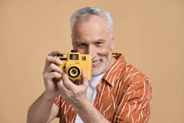 英俊的白人老人 穿着橙色休闲服的游客 手持明亮的黄色相机 在拍照时面带微笑 与米色背景隔离 人与夏天的概念 — 图库照片