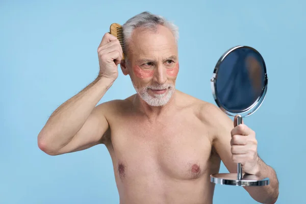 Attractive seductive grey-haired bearded senior man with hydrogel patches under eyes, combing hair with wooden comb, smiles looking at his reflection in a cosmetic mirror, isolated on blue background