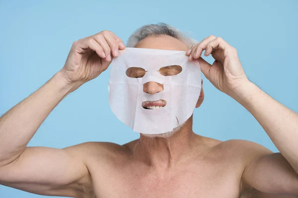 Male beauty and skin care concept. Caucasian senior man putting a hydrating purifying tissue mask on his face, standing with naked torso isolated over blue color background